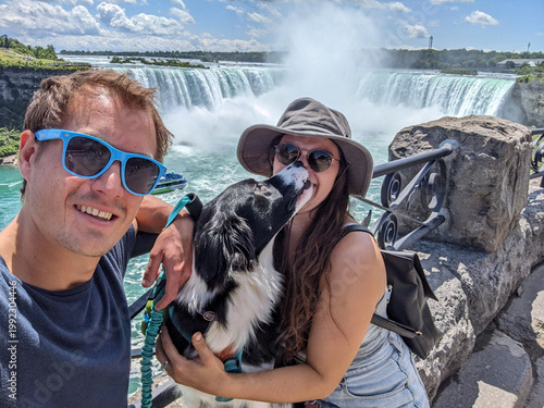 Happy couple pose with their dog for a selfie at Niagara Falls. The border collie kisses the woman while enjoying sunny weather and misty waterfall views on a scenic riverside overlook.