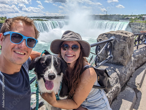 A young couple and their dog smile for a selfie with Niagara Falls in the background during a sunny day in the summer.