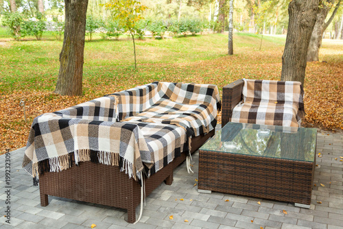 Outdoor wicker sofa, armchair, and glass table with plaid blankets on a paved patio. Autumn setting with fallen brown leaves and trees in a park.