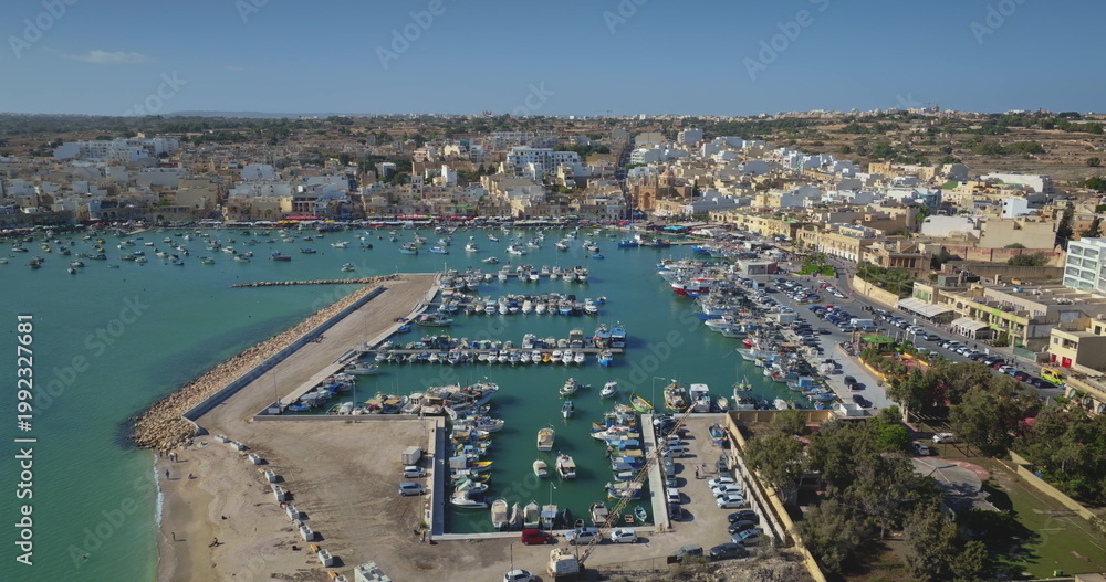 Obraz premium Malta: Aerial view of Marsaxlokk harbor, featuring colorful traditional Luzzu boats anchored in the port on a sunny summer day. Drone flight