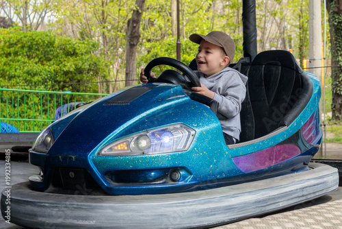 a boy laughs openly with great joy while steering a blue and purple glitter bumper car with illuminated headlights against a backdrop of lush green spring trees
