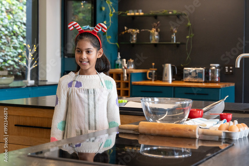 Indian girl child wearing antler headband and apron mixing batter at kitchen island with glass bowl