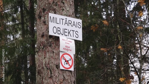 Military object warning sign displayed on a tree in a forested area, emphasizing restricted access and drone prohibition