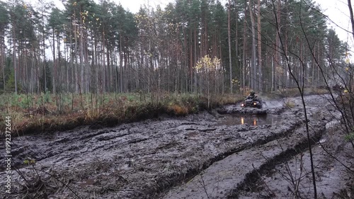 Six-wheeled ATV navigating muddy terrain in a forest, splashing water and mud while maneuvering through challenging conditions