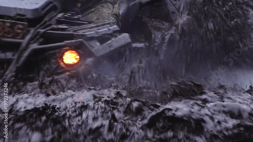 Six-wheeled ATV splashes through muddy water, creating spray and mist while navigating a rugged terrain in a forested environment