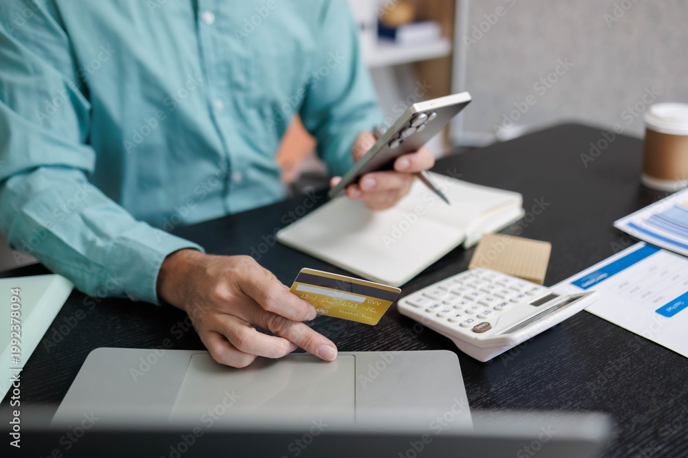 Fototapeta premium A person is holding a credit card and a pen on a desk. The desk has a calculator, a cell phone, and a laptop. The person appears to be working on a financial transaction or a budget.