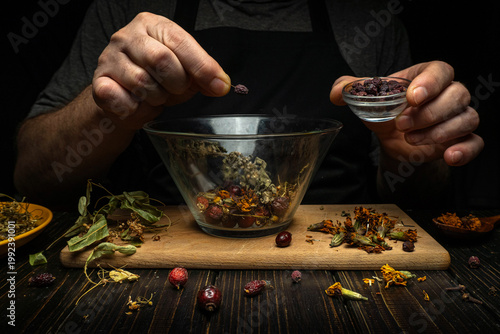 A person adds dried herbs and flowers to a glass bowl on a wooden board. They hold a small dish in their other hand. The kitchen is dimly lit, creating a focused scene