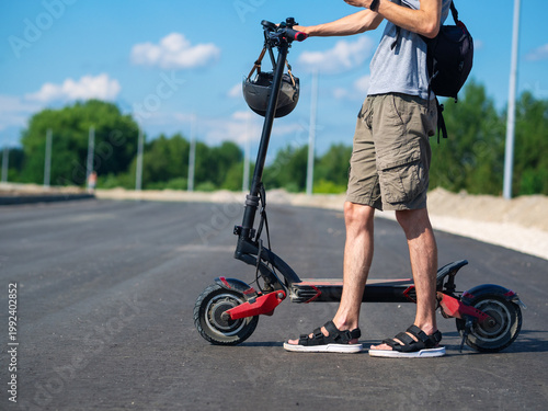A young man with a backpack stands by an electric scooter on the street. A helmet for traveling. summer urban transport