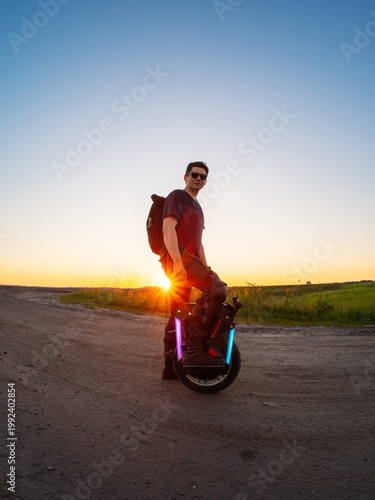 A man poses standing on a mono wheel, a summer sunset and a country road. modern electric individual transport.