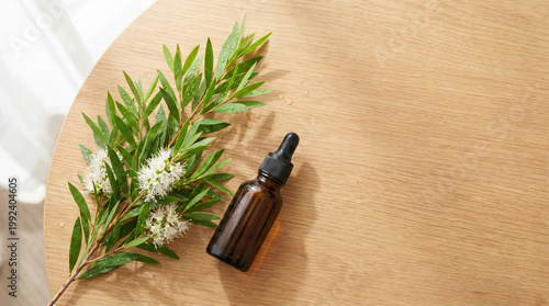 A dark amber glass dropper bottle rests on a light wooden table beside a fresh green branch of a blooming tea tree with white flowers and water droplets in soft sunlight.