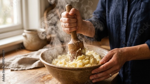 Hot, creamy mashed potatoes are being prepared with a wooden masher in a cozy kitchen. Steam rises from the bowl.
