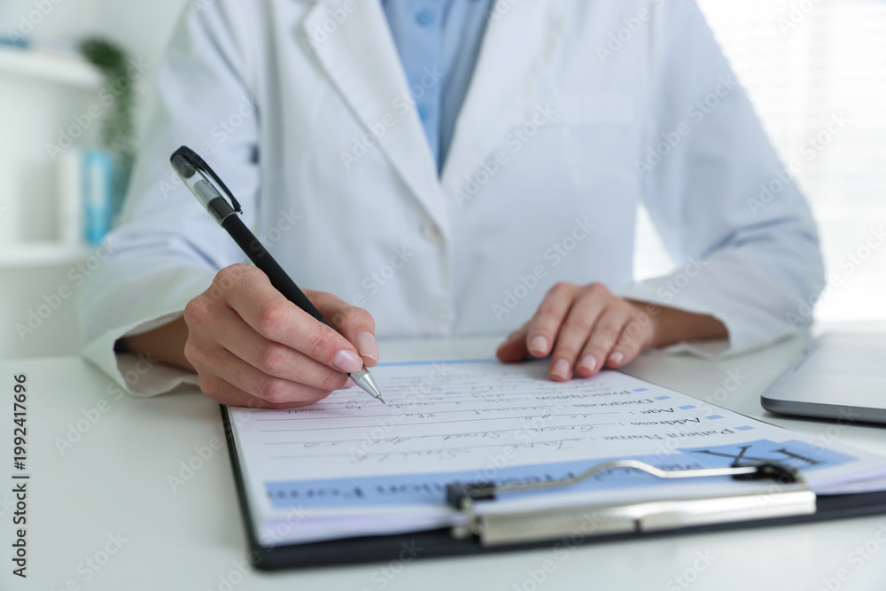 Fototapeta premium Doctor in medical coat writing on clipboard at white table in clinic, closeup