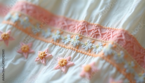 Close view of delicate smock embroidery on creamy white cotton fabric. Pastel threads create floral motifs and decorative patterns. Soft texture and intricate stitching show fine craftsmanship.