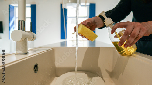 Hands holding sponge and dish soap under running water in a modern kitchen sink