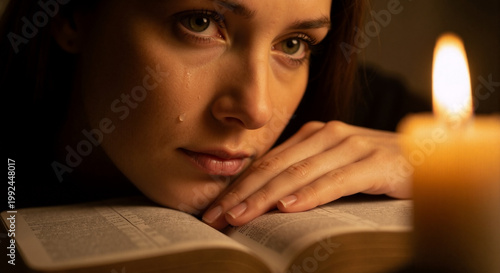 Close up of a young woman with a tear on her cheek resting on an open Bible. Spiritual grief and religious faith concept. Candlelight illuminating a tearful female face reading scripture