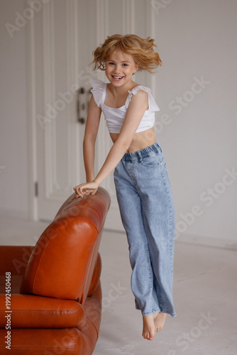 Smiling child in white sleeveless top and denim shorts posing behind orange leather sofa in bright living room, front view, childhood, family lifestyle, happiness, advertising concept. 