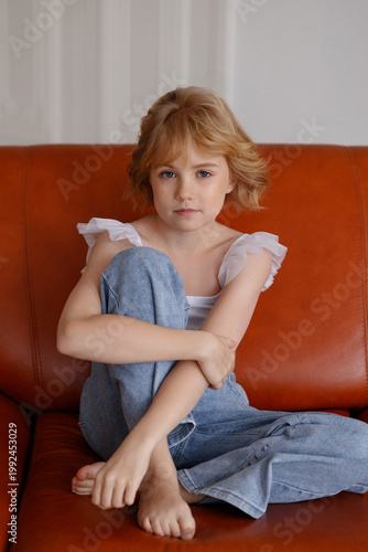 Young girl, fair skin, blonde hair, casual clothes, sitting with legs crossed on orange leather sofa, front view, look at camera, bright room. Concept of childhood, home life, calm mood.