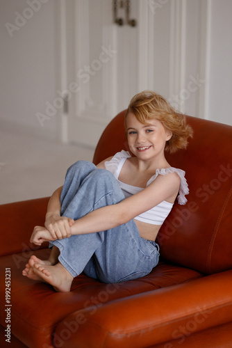 Young Caucasian girl, child, blonde hair, casual clothes, sitting with legs folded, smiling, look at camera, brown leather armchair, white room. Concept childhood, comfort, home, lifestyle.