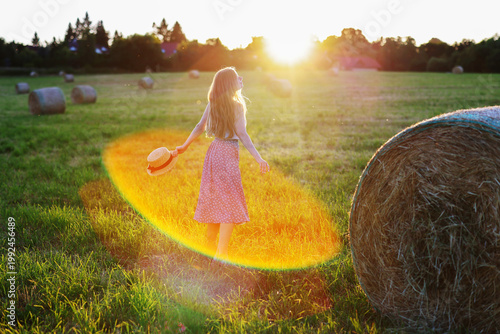 Woman walking in sunset field with hat in hand, dreamy rural summer evening light.
