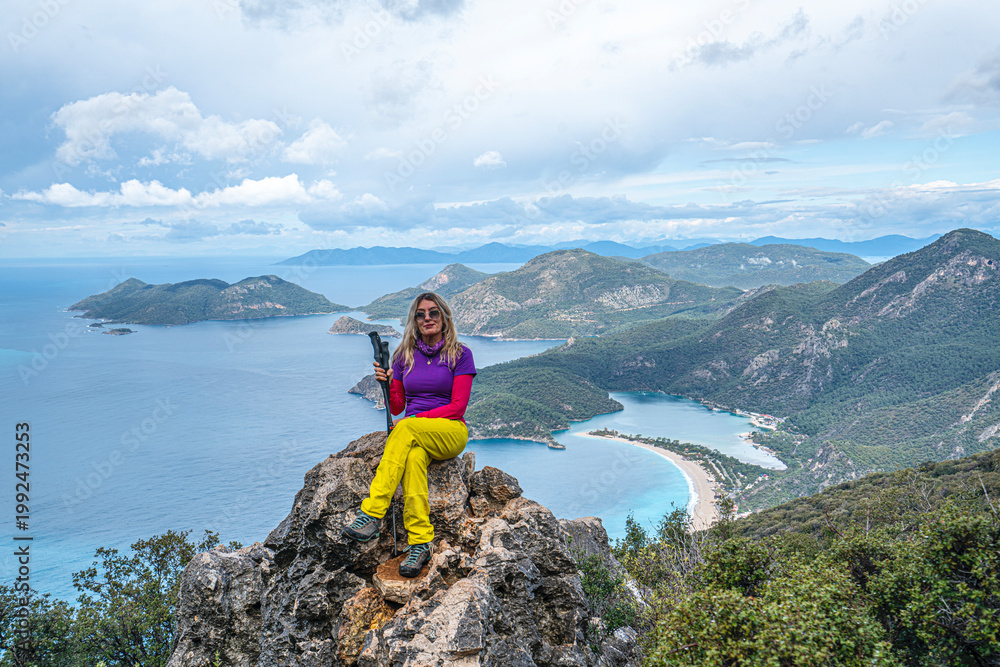 Fototapeta premium Beautiful blond woman hiker is enjoying the view of Ölüdeniz, which is a resort village on the southwest coast of Turkey, known for the blue lagoon and the wide, white Belcekız Beach. On the route of 