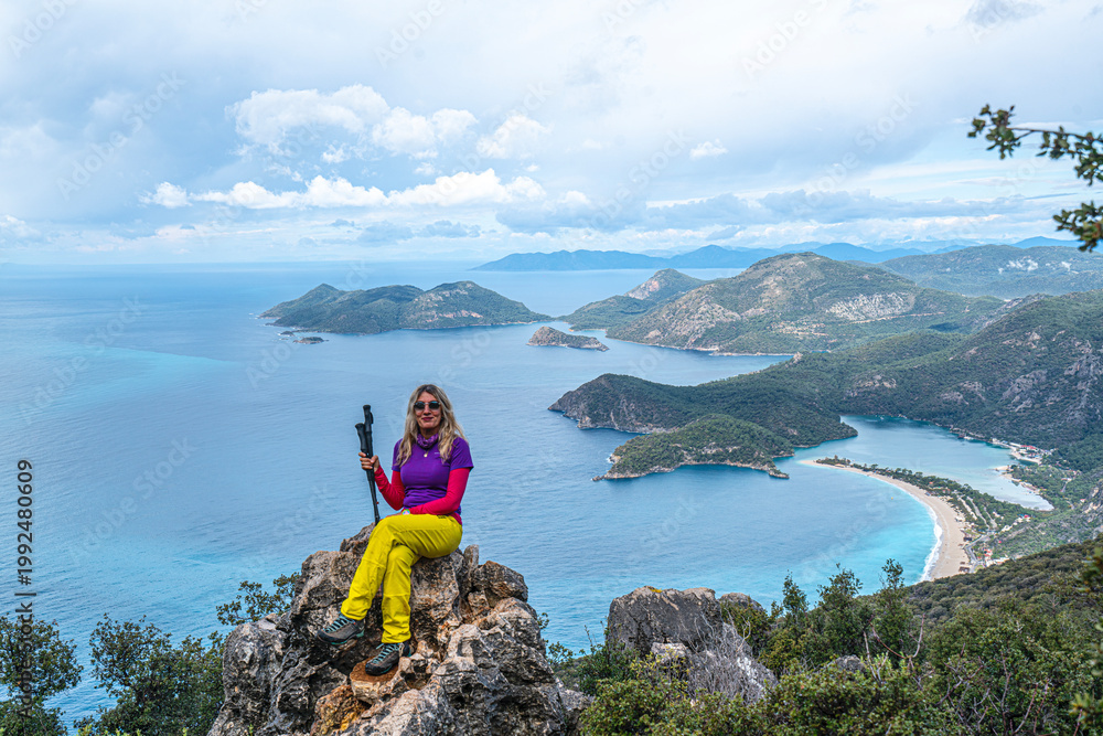 Fototapeta premium Beautiful blond woman hiker is enjoying the view of Ölüdeniz, which is a resort village on the southwest coast of Turkey, known for the blue lagoon and the wide, white Belcekız Beach. On the route of 
