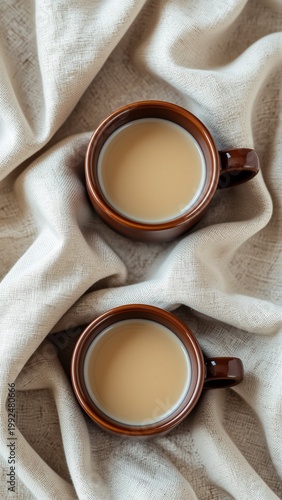 two brown ceramic mugs filled light colored liquid which appears tea coffee mugs placed beige fabric crumpled texture background blurred making mugs focal point taken top down perspective focus cups