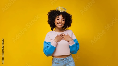 Happy young woman in casual trendy outfit with crown against yellow background expressing joy and surprise lifestyle
