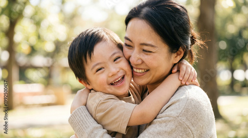 Smiling Asian mother and her young son with Down syndrome hugging in a park, inclusive family life and outdoor joy.