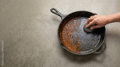 Top-down image of a rusted cast iron skillet being cleaned with a woman’s hand holding a stainless steel scouring pad. Copy space. Minimalist composition.