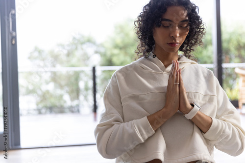 Non-binary person pressing palms at chest near sliding doors, wearing hoodie, smartwatch and ring