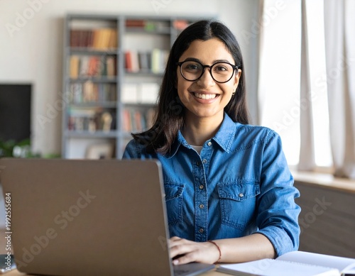 Portrait of happy young woman or student wearing glasses sitting at desk at home office with laptop computer looking at camera advertising online learning, remote work, business webinars