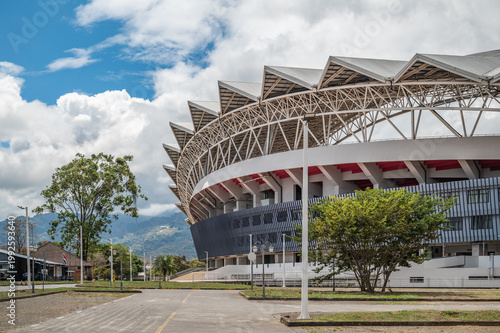 Modern National Stadium in La Sabana Park, San Jose, Costa Rica, iconic sports venue with steel architecture, surrounded by trees, city streets, and mountains under bright cloudy sky (February 2026).