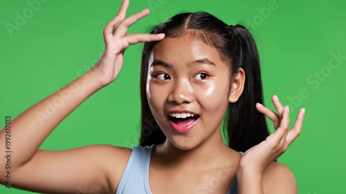 Young Asian girl with pigtails applying moisturizer, smiling on green screen
