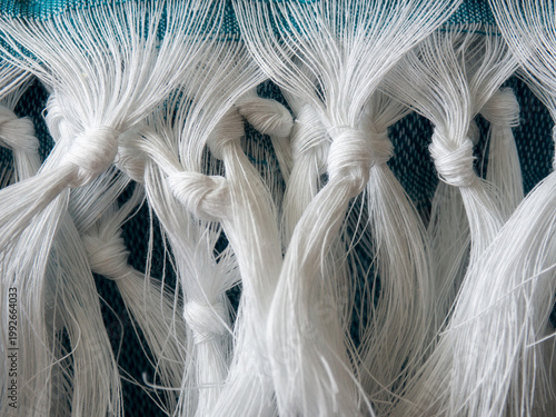 Macro close-up of white knotted fringe threads on blue textile fabric. Detailed woven texture and decorative fiber pattern