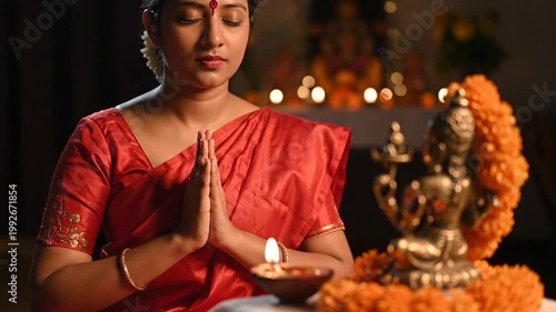 Indian woman in red sari praying with hands joined in namaste, with a lit diya lamp and Hindu deity idols decorated with marigold flowers, creating a peaceful devotional atmosphere