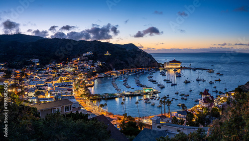 Coastal town at dusk with a curved harbor full of boats, warm building lights, surrounding hills, and a prominent round waterfront landmark.
