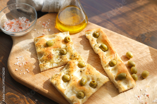 Traditional Italian olive focaccia slices on wooden board with pink salt and olive oil. High-angle shot captures authentic Mediterranean bakery texture and rustic homemade aesthetic under warm natural