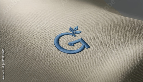 Embroidered letter G logo on white fabric texture, blue thread embroidery on cotton textile, elegant monogram branding mockup for fashion apparel