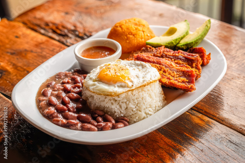 A traditional Colombian bandeja paisa served on a white plate, featuring white rice topped with a fried egg, red beans, chicharron, arepa, avocado slices, and a small cup of hogao sauce.