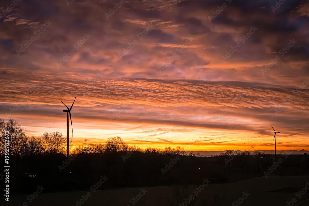 Obraz premium Die Nacht zieht sich langsam zurück wie ein schwerer Vorhang, und dahinter glüht der Himmel in flüssigem Gold und flammendem Kupfer. Die Windräder stehen wie stille Wächter am Horizont, ihre Flügel sc