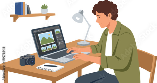 A man sits at a desk working on a laptop with a camera and notebook nearby.