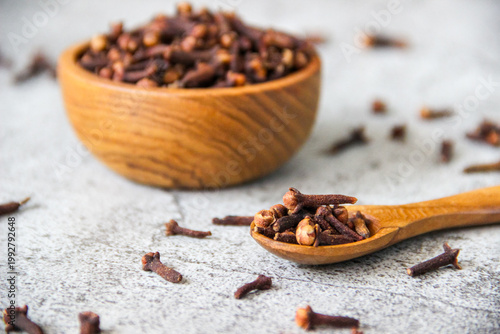 Dark brown cloves fill a wooden bowl. Scattered cloves rest on a gray concrete table. Wooden spoon holds a few cloves