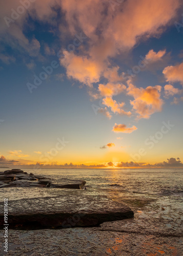 A sunrise breaking through a cloud bank lights up the clouds with its orange glow and reflects in the water on a rock platform jutting out to sea at Burrill Lake in New South Wales, Australia.