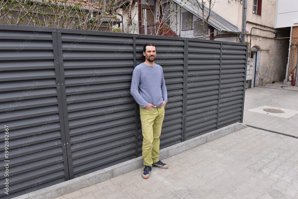 Fototapeta premium Man in grey sweater and yellow pants leaning against a dark louvered metal privacy fence