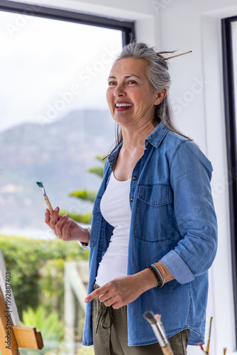 Senior woman standing in bright art studio holding paintbrush with green paint, denim shirt