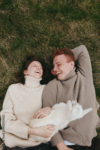 Fotografia Joyful couple lying on grass with a fluffy white cat, warm sweaters, relaxed moo