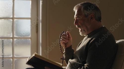 Mature man in quiet prayer, holding a wooden cross rosary and whispering words from the Bible by a sunlit window