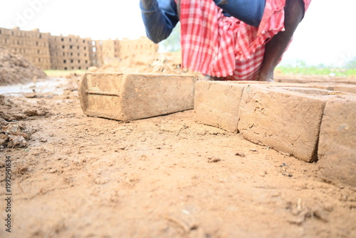 A person manually shaping bricks in an open field. Using a rectangular wooden mold and raw, reddish-brown clay, the worker skillfully forms each brick on the dusty ground. Slow motion video.