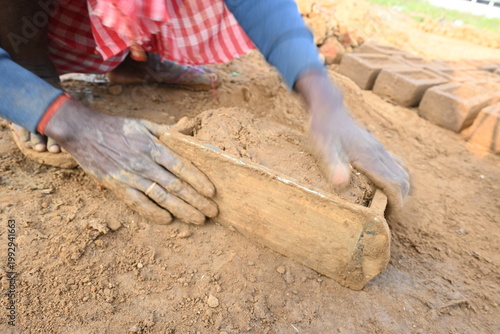 A person manually shaping bricks in an open field. Using a rectangular wooden mold and raw, reddish-brown clay, the worker skillfully forms each brick on the dusty ground. Slow motion video.