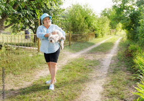 Happy senior woman carrying small white dog in summer garden.